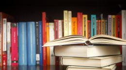 Stack of books on a wooden library shelf, the one on top open , multicolored book spines in a row in the background. Copy space on the left.
